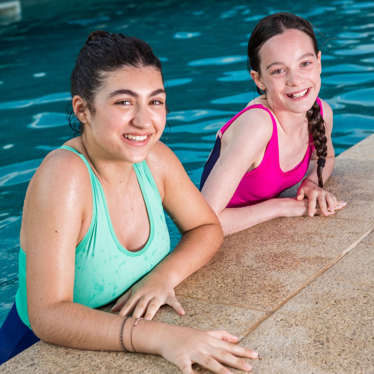Two teenage girls wearing WUKA Teen Period Swimsuits in Green/Blue and Pink/Purple, smiling and relaxing at a swimming pool edge.