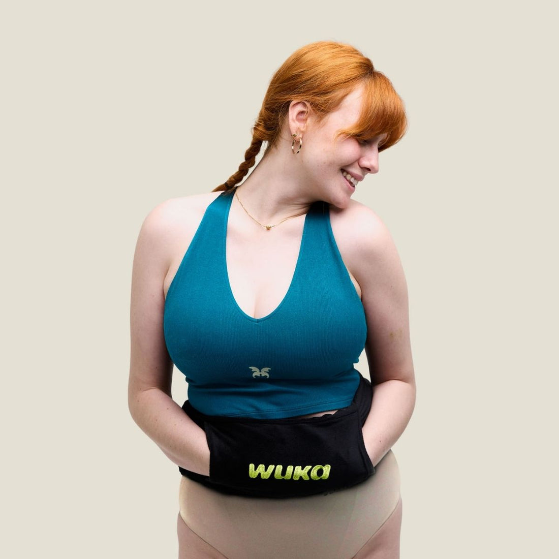 Smiling red-haired woman in a teal crop top and period pants holding a black WUKA hot water bottle against her stomach. She’s standing confidently against a neutral background, radiating comfort, confidence, and period pride.