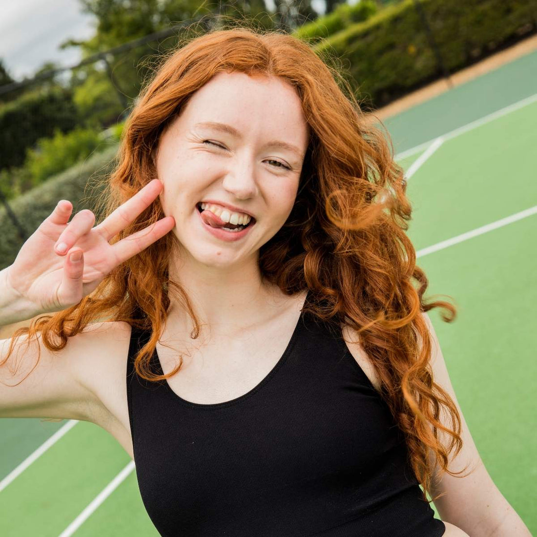 Teen winking and flashing a peace sign on a tennis court - proof that periods don’t pause play. Moving your body your way, period or not.