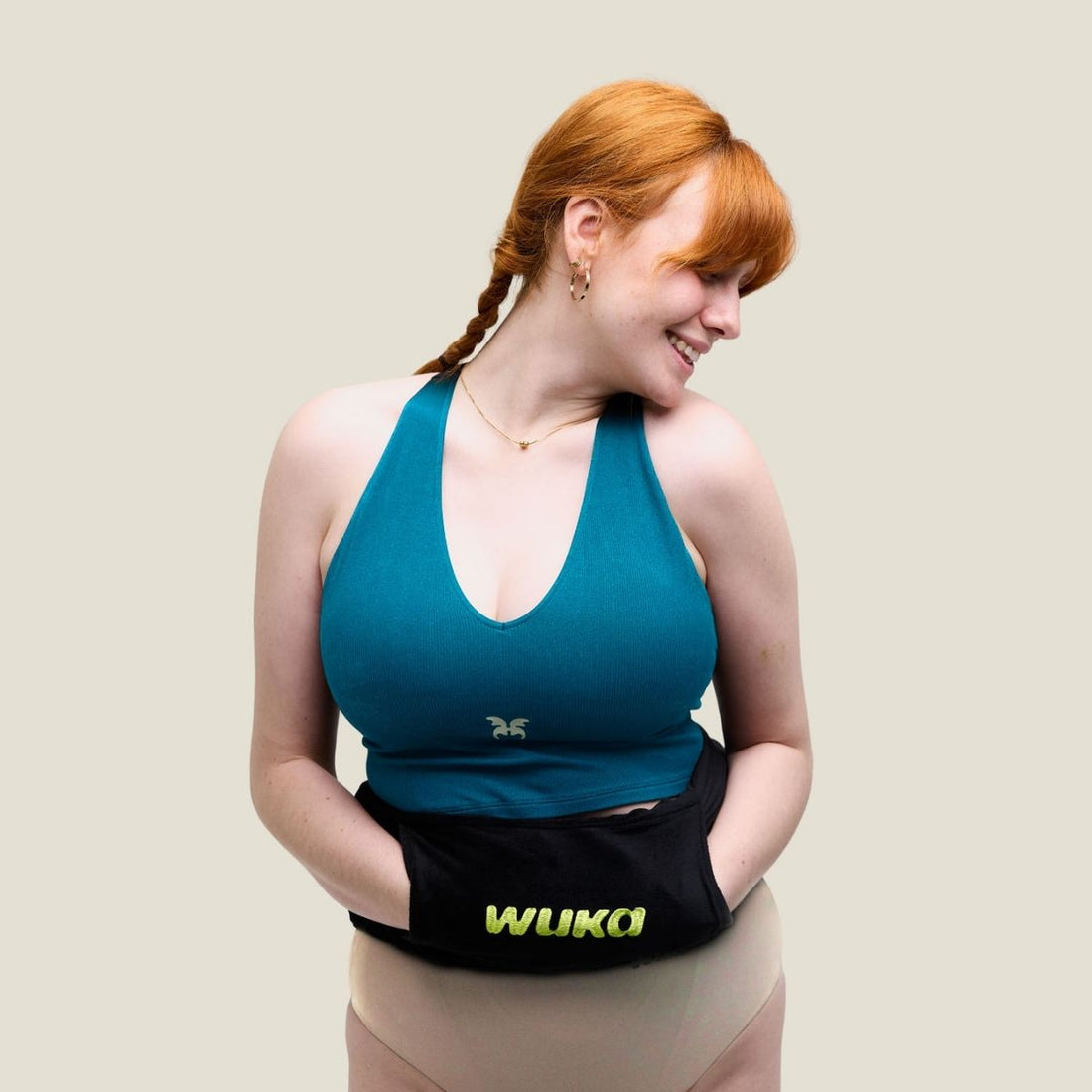 Smiling woman with red hair wears a teal sports top and WUKA wearable hot water bottle around her waist, standing confidently against a neutral background.